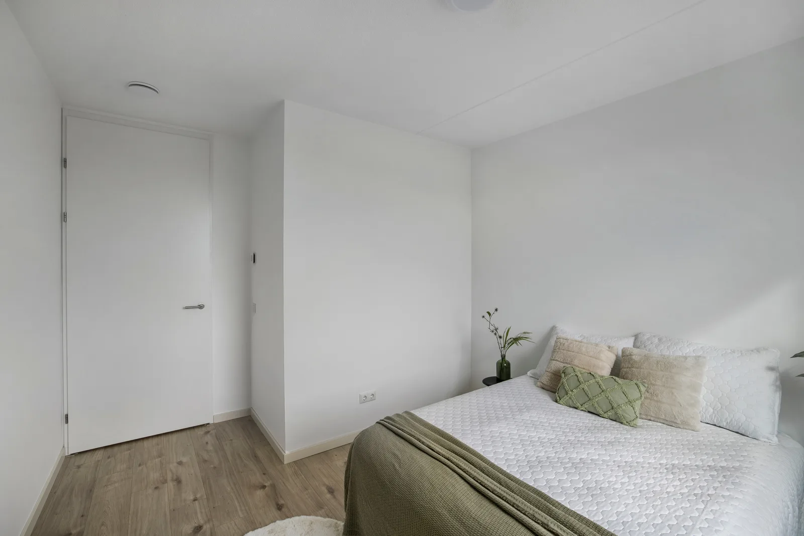 Slaapkamer met witte wanden, lichte houtlook vloer, opgemaakt bed rechts en een witte binnendeur