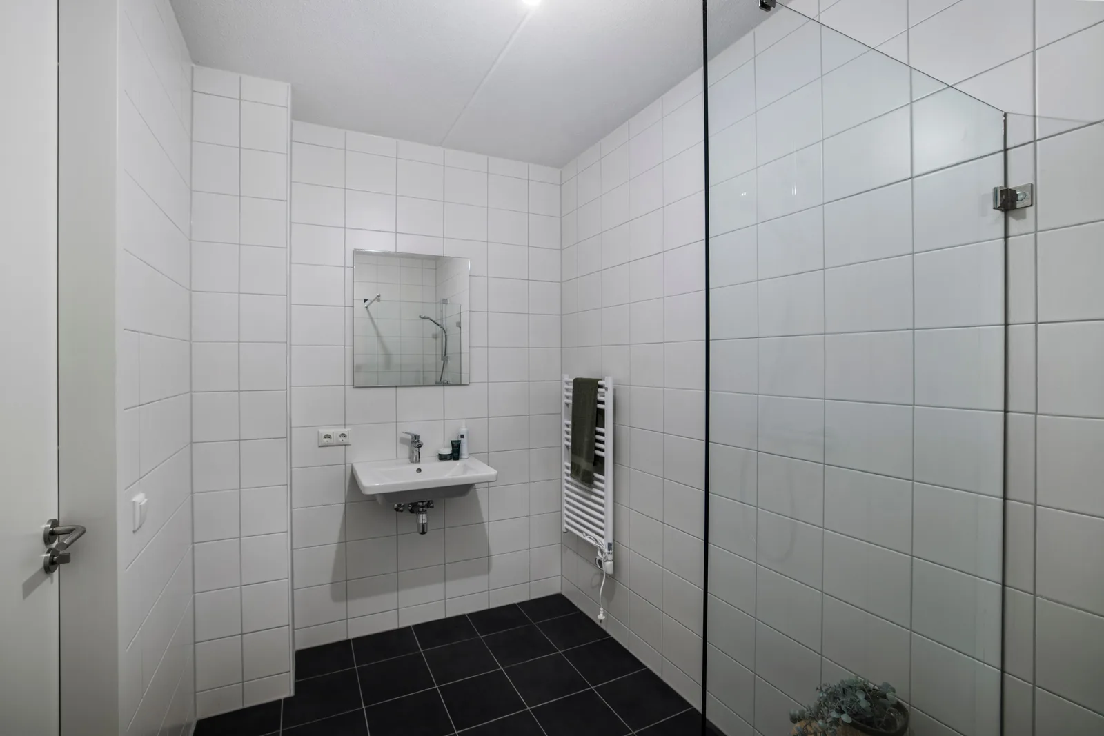 Badkamer met witte wandtegels, antracietgrijze vloertegels, hangende wastafel met spiegel en rechts een glazen douchewand