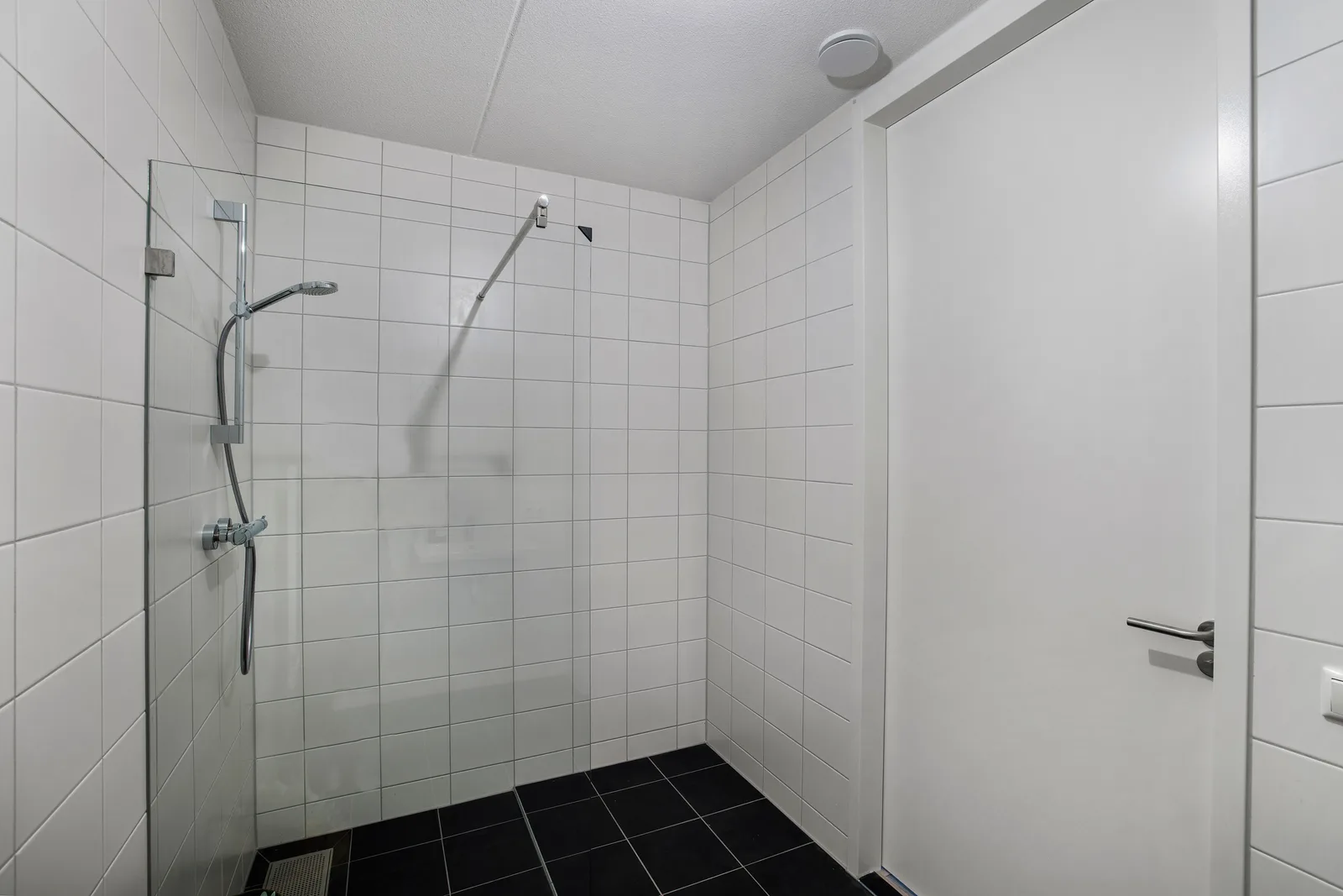 Inloopdouche met chromen doucheset achter een glazen wand, witte wandtegels en zwarte vloertegels