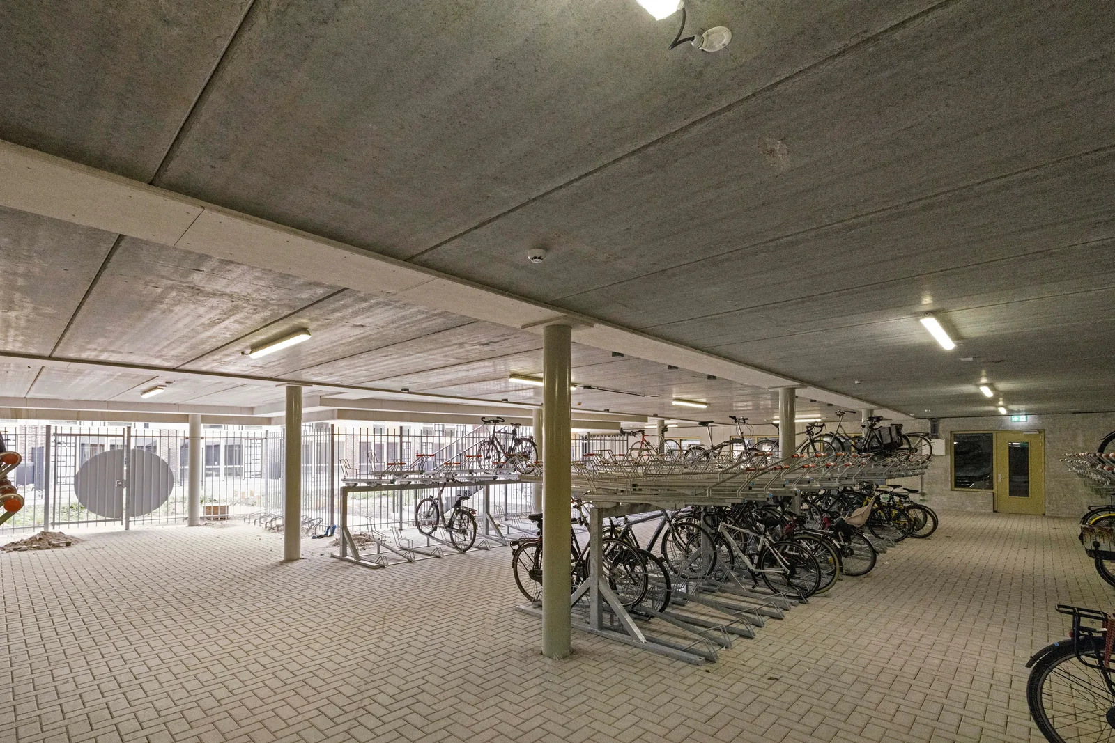 Overdekte fietsenstalling met betonnen plafond, ronde kolommen, dubbellaagse fietsenrekken en rechtsachter een gele dubbele deur
