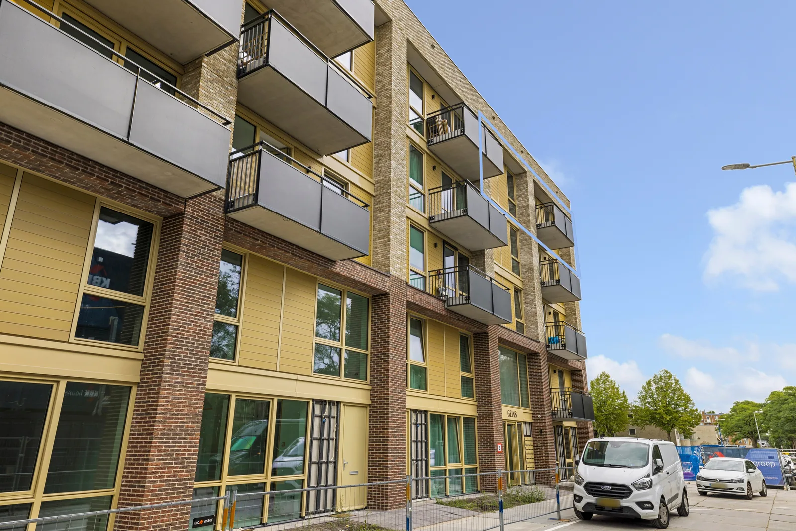 Appartementengebouw met gele gevelpanelen, roodbruine en gele baksteen, donkergrijze balkons en grote ramen beneden