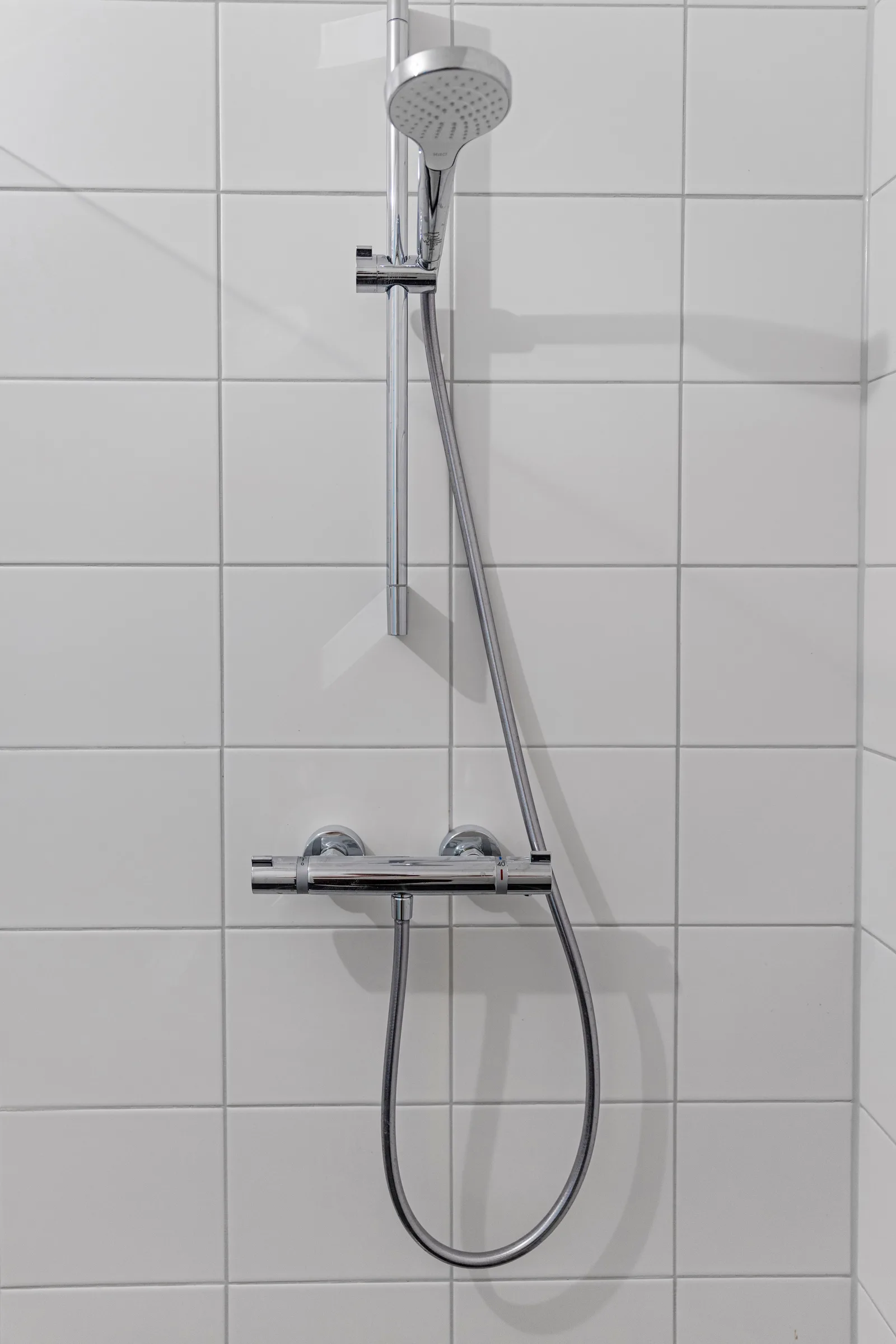 Chromen thermostaatkraan met handdouche aan een verticale glijstang tegen witte wandtegels
